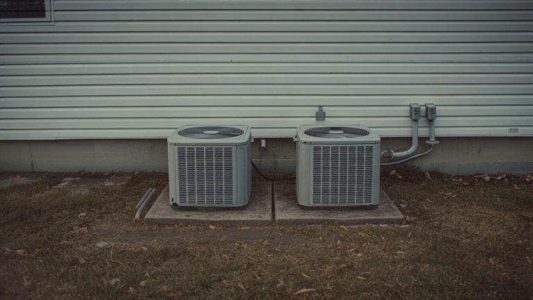 Two residential air conditioning units sitting on a concrete pad beside a house on a mild off-season day